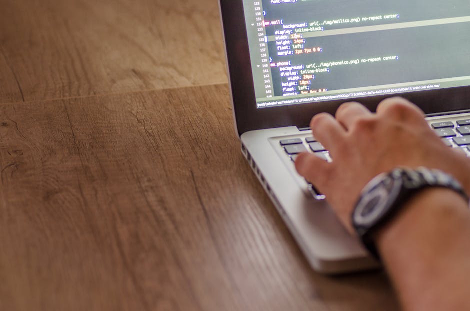 A close-up view of a person using a laptop computer on a wooden surface, with the individual's right hand resting on the keyboard. The person is wearing a wristwatch with a dark strap and a metallic w