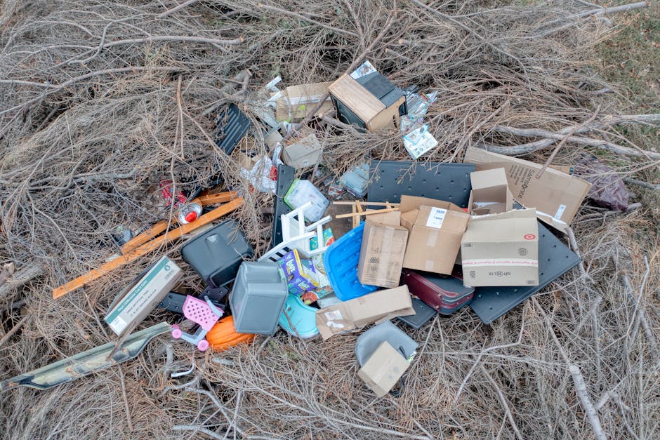 A pile of miscellaneous household waste and discarded items accumulated on a patch of dry, brown grass and leafless twigs outdoors. The collection includes various plastic containers, such as a blue l